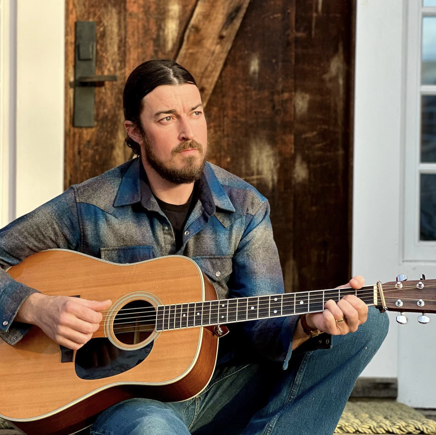 Man in denim jacket playing acoustic guitar outside.