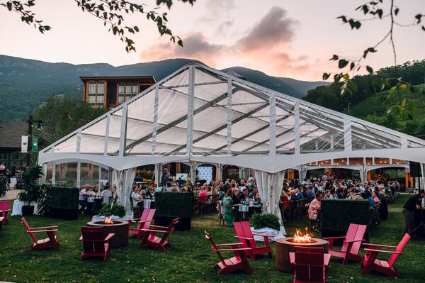 Outdoor gathering under a white tent with people, chairs, and a fire pit at sunset.