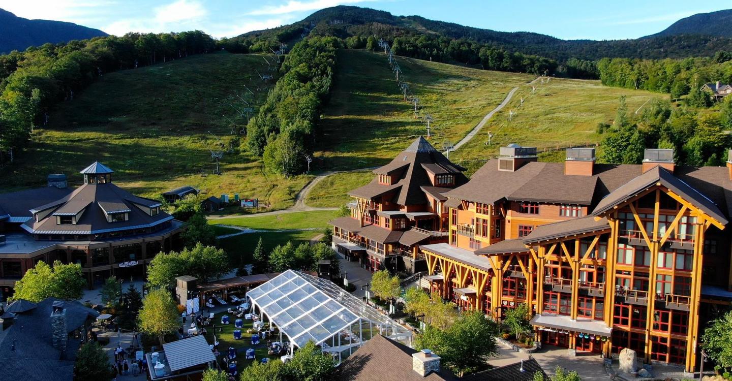 Lodge with wooden architecture near a grassy hillside, surrounded by mountains.