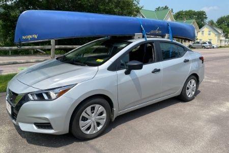 Silver car with a blue canoe on top, parked on a sunny street.