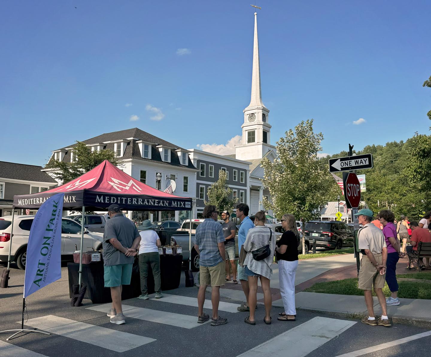 Street fair with food stall, people gathering, church steeple in the background.