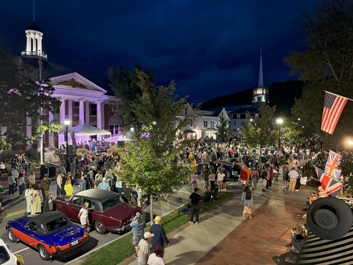 Crowd gathered at night in a town square with classic cars and a lit-up historic building.