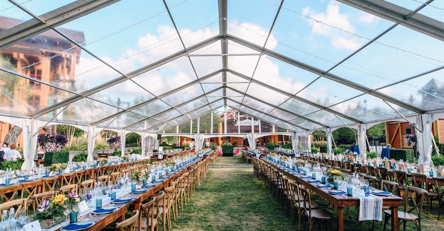 Clear tent setup with long banquet tables and chairs on grass, under blue sky.