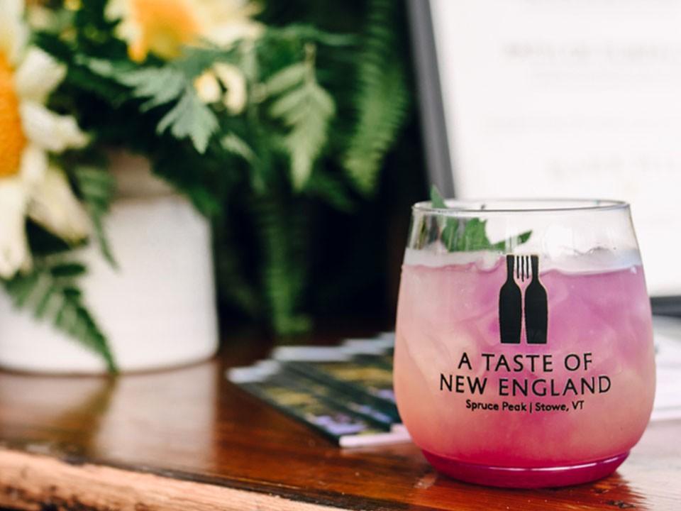 Pink cocktail glass with "A Taste of New England" logo, on a wooden table with flowers nearby.