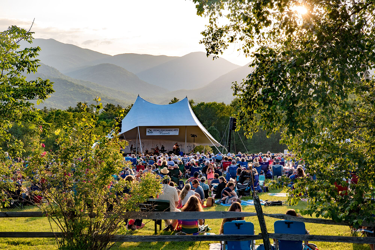 Outdoor concert with a crowd, tent stage, and mountains at sunset.