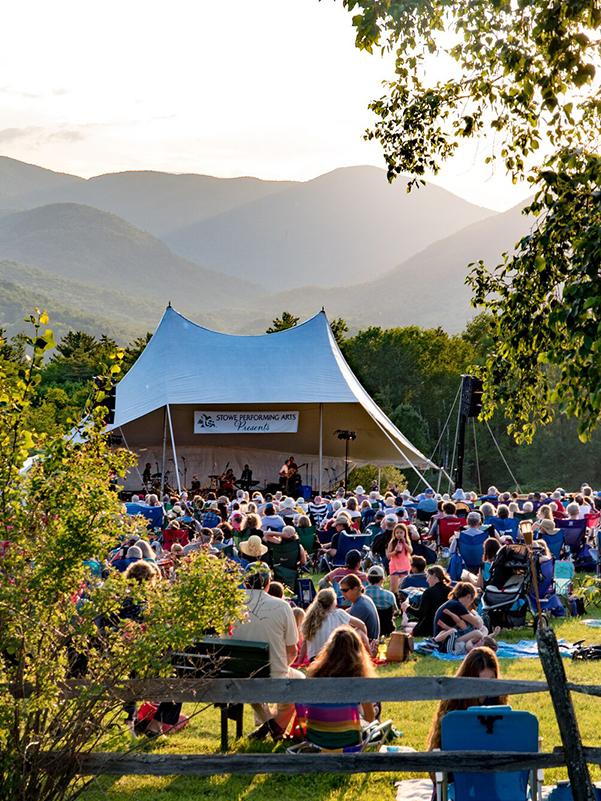 Outdoor concert with crowd seated on grass, tent stage, and mountains in the background.