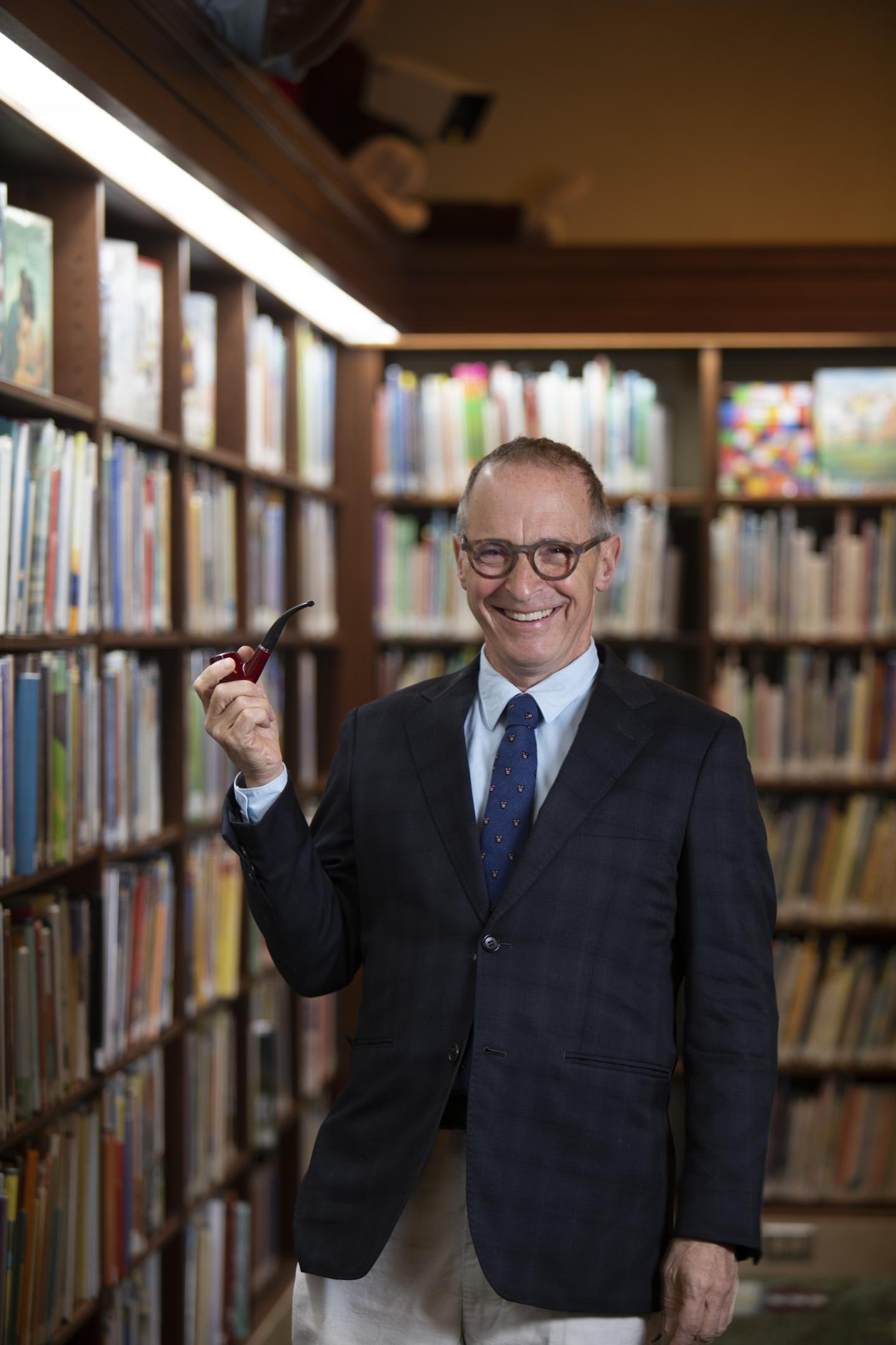 Man in a library holding a red object, wearing glasses and a suit.