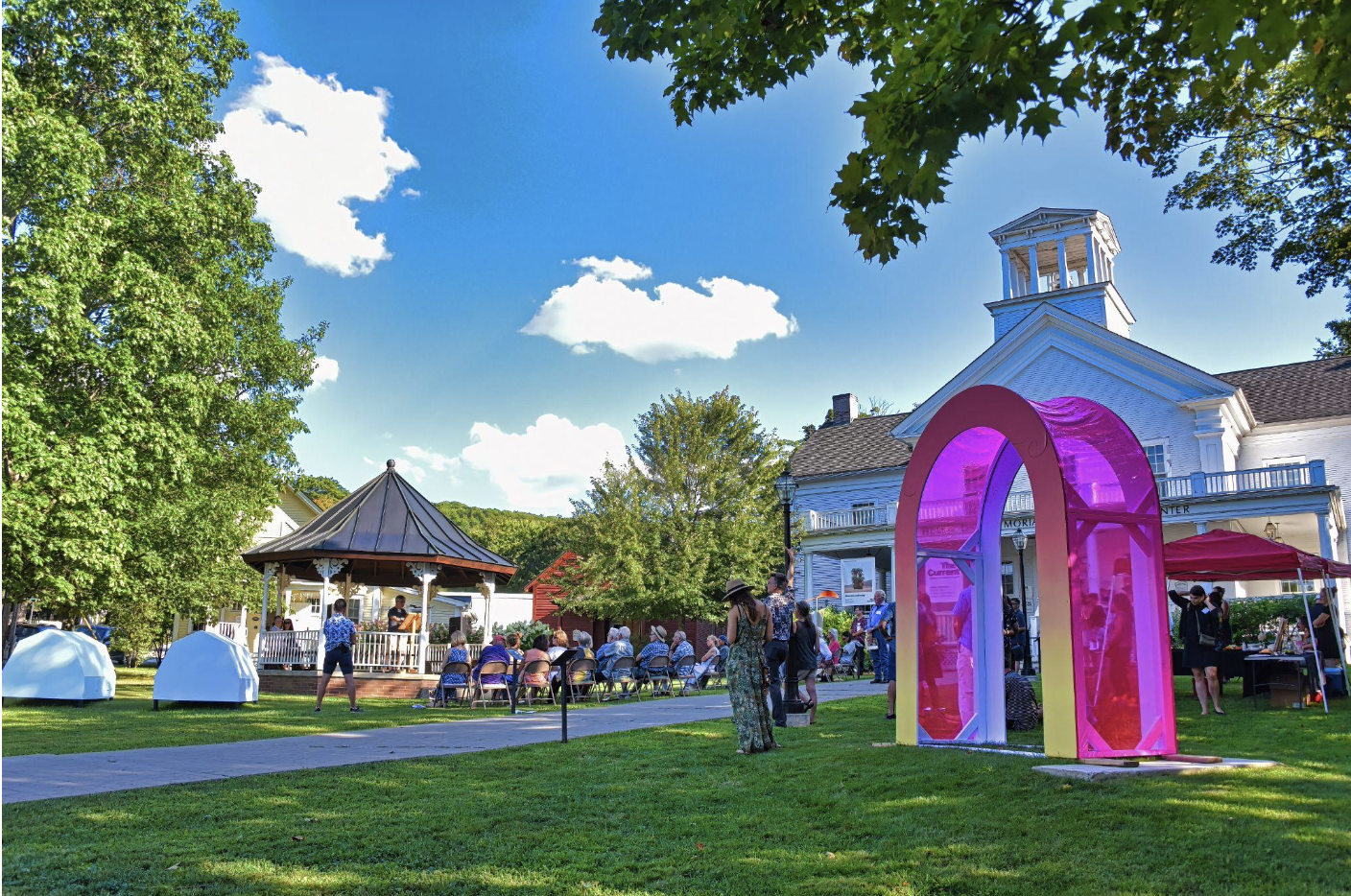 Sunny outdoor event with a gazebo, colorful arch, and a crowd near a white building.