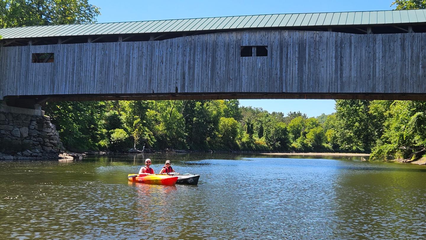 Two kayakers on a river beneath an old wooden covered bridge.