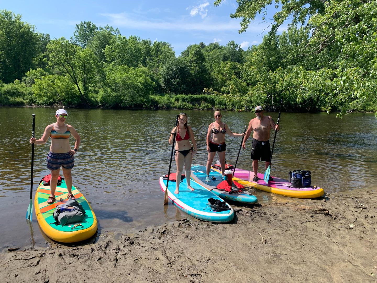 Four people with paddleboards on a riverbank, surrounded by lush greenery.