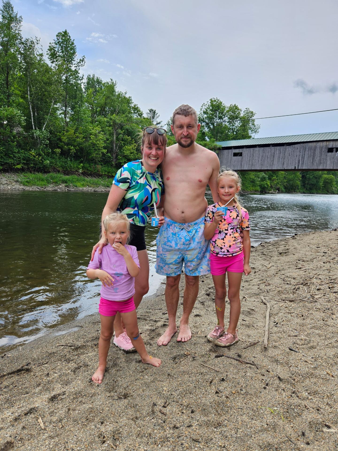 Family of four standing on a sandy riverbank with a wooden bridge in the background.
