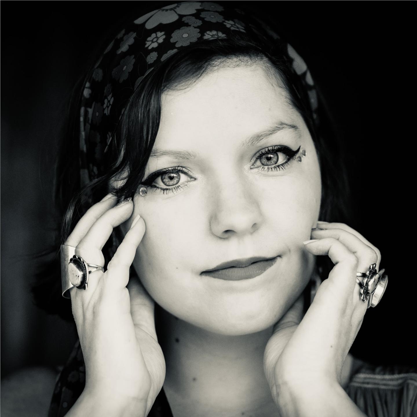 Black and white photo of a woman with a headscarf, eyeliner, and rings, touching her face.
