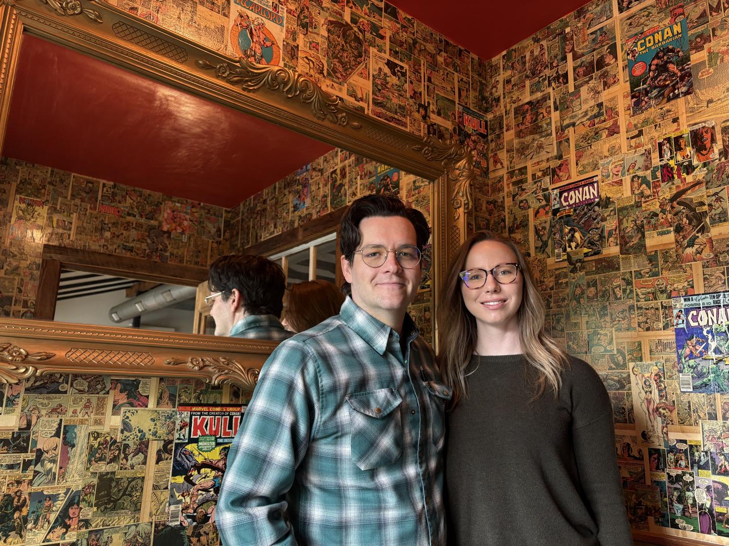 Two people smiling in a room with comic-covered walls.