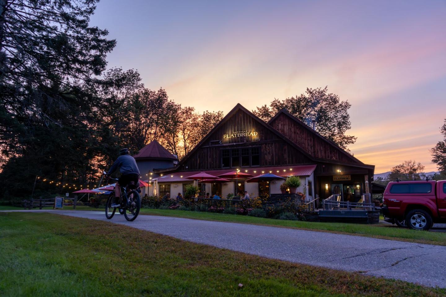 Cyclist rides past rustic building at sunset, with lit patio and trees.