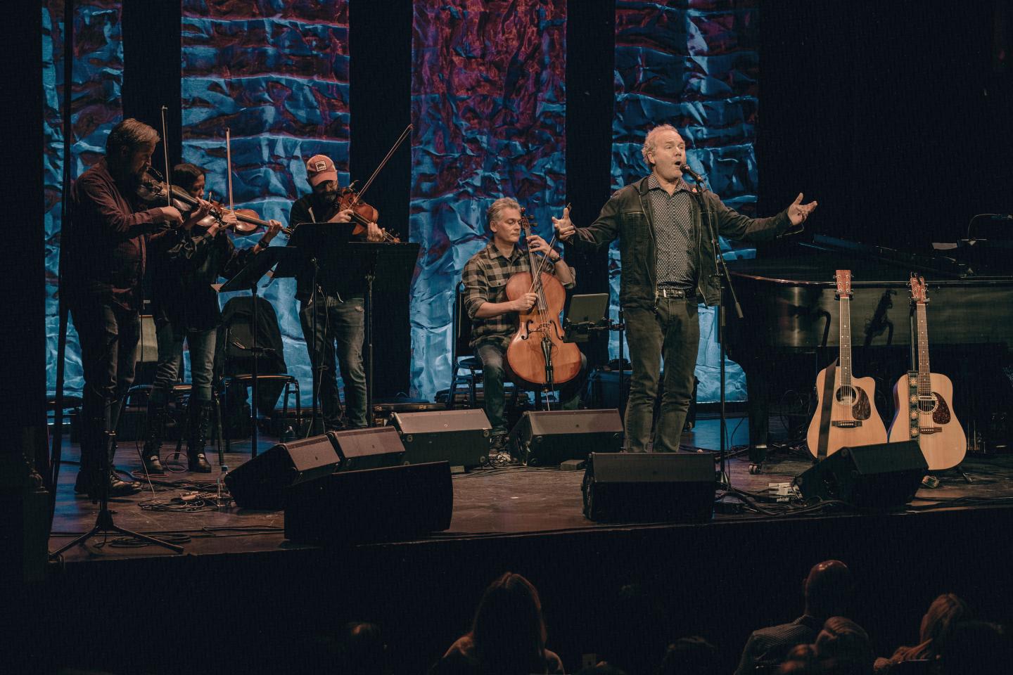 Musicians perform on stage with violins and cello under colorful lights.