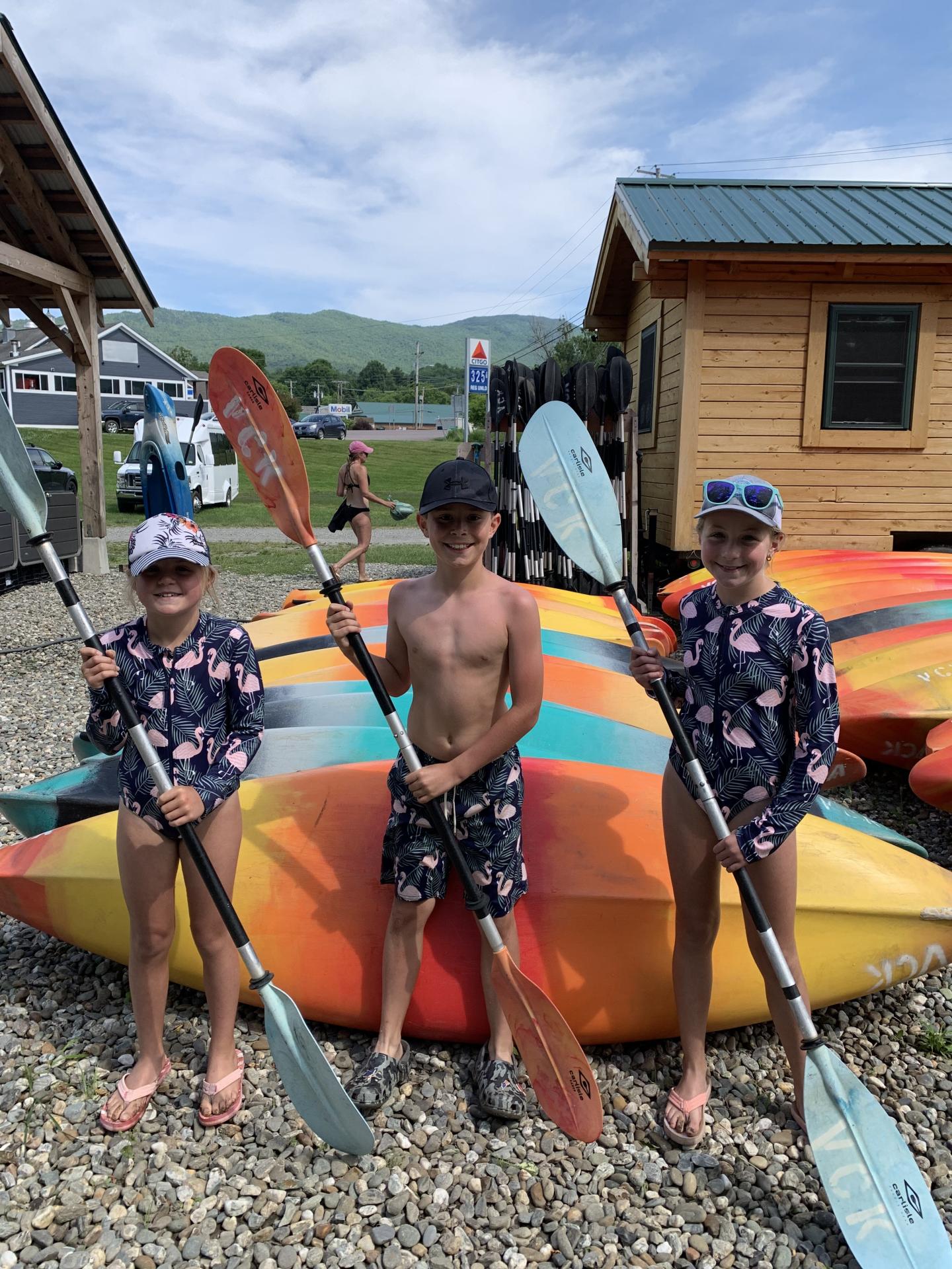 Children holding paddles, standing in front of colorful kayaks.