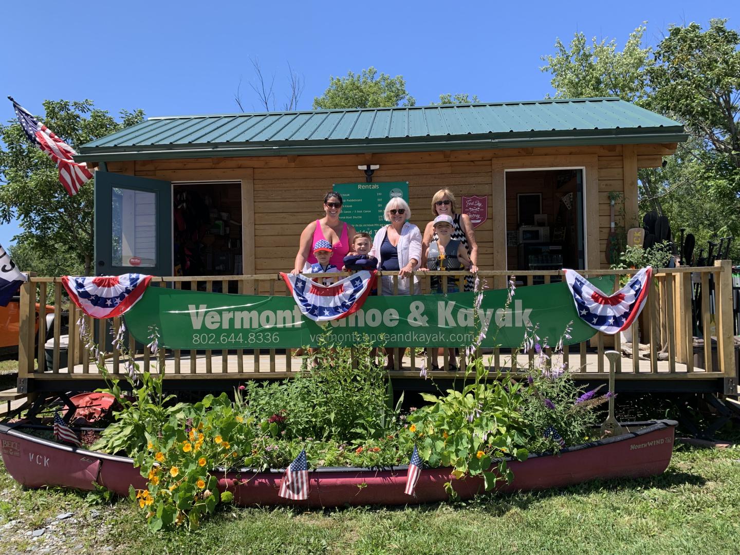 Three people stand on a deck in front of a wooden shack with a green sign and festive flags.