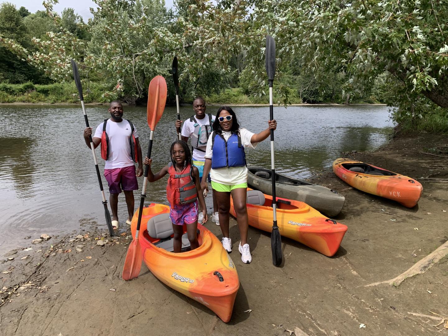 Family with kayaks by a lake, holding paddles, surrounded by trees.
