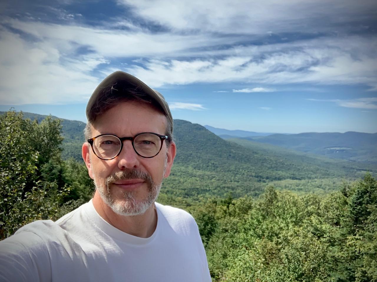 Man in glasses and cap smiling with a scenic mountain view background.