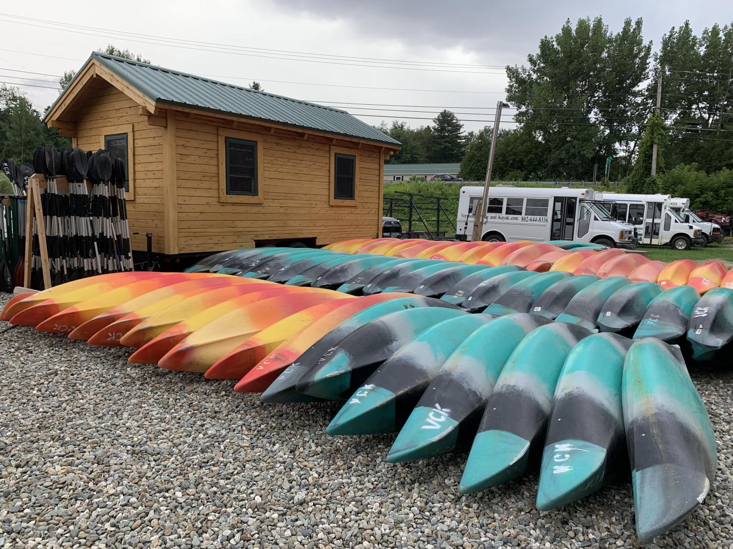 Colorful kayaks lined up by a wooden shed and white vans.