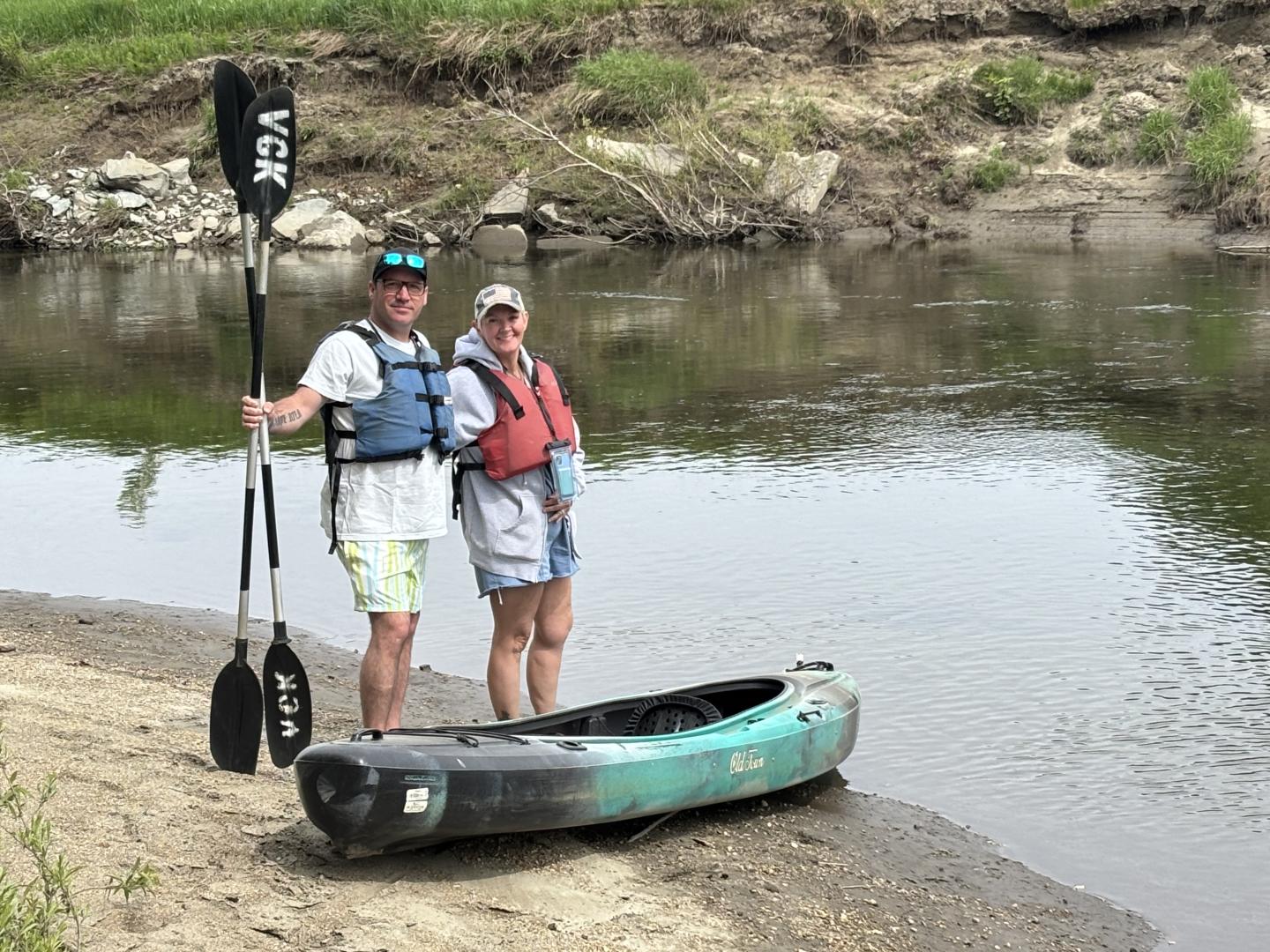 Two people in life jackets stand by a green kayak on a riverbank.