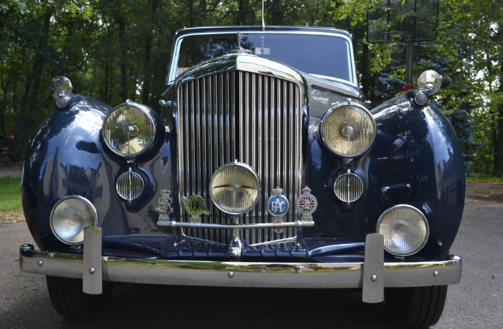 Vintage blue luxury car with chrome grille and headlights in a forested area.