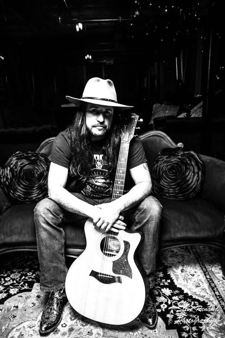 Man with guitar, wearing a hat, sitting on a sofa in a dark room. Black and white photo.