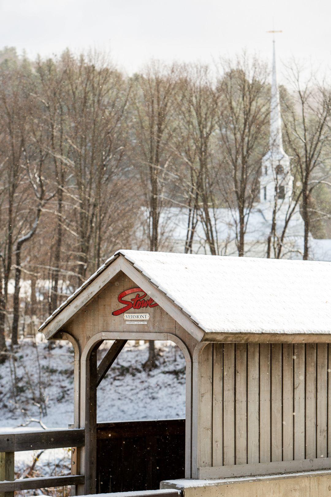Snow-covered bridge and trees with a distant church spire.