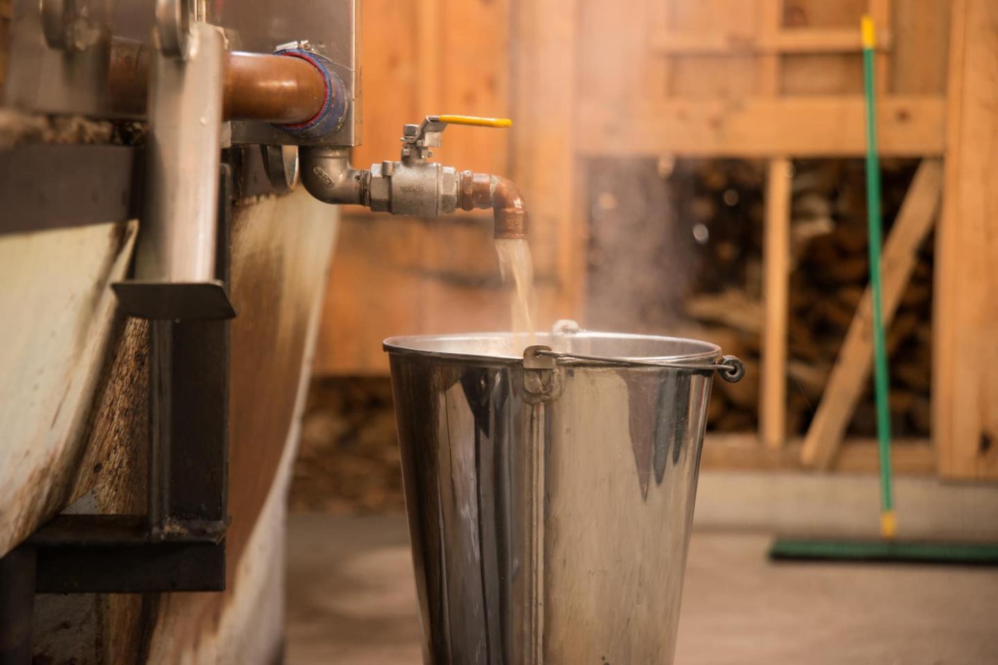 Steam rises from hot liquid pouring into a metal bucket in a wooden room.
