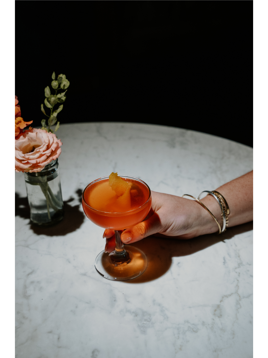 Hand holding an orange cocktail on a marble table, flowers in the background.