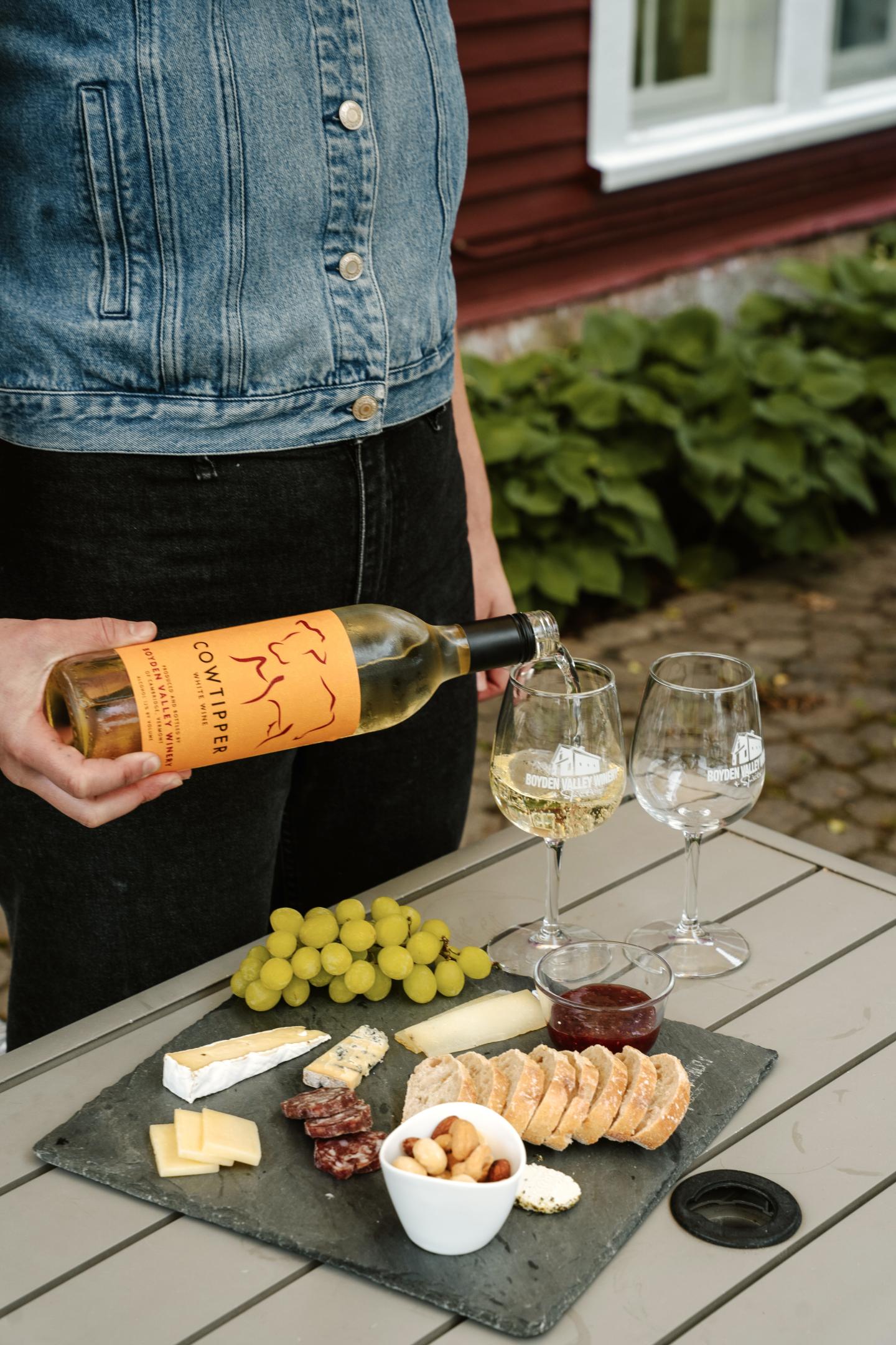 Woman holding wine bottle over outdoor cheese platter with grapes and glasses.