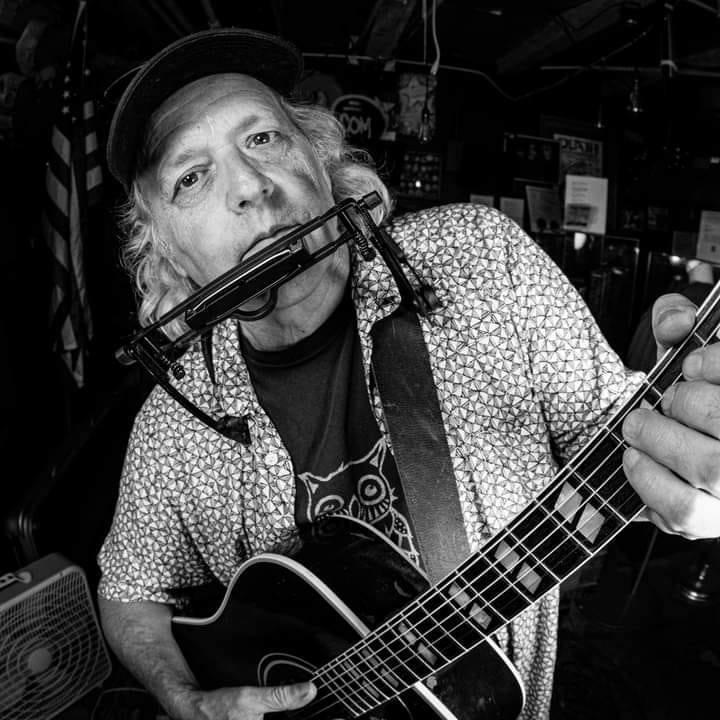 Man playing guitar and harmonica, wearing a cap, in a dimly lit room.