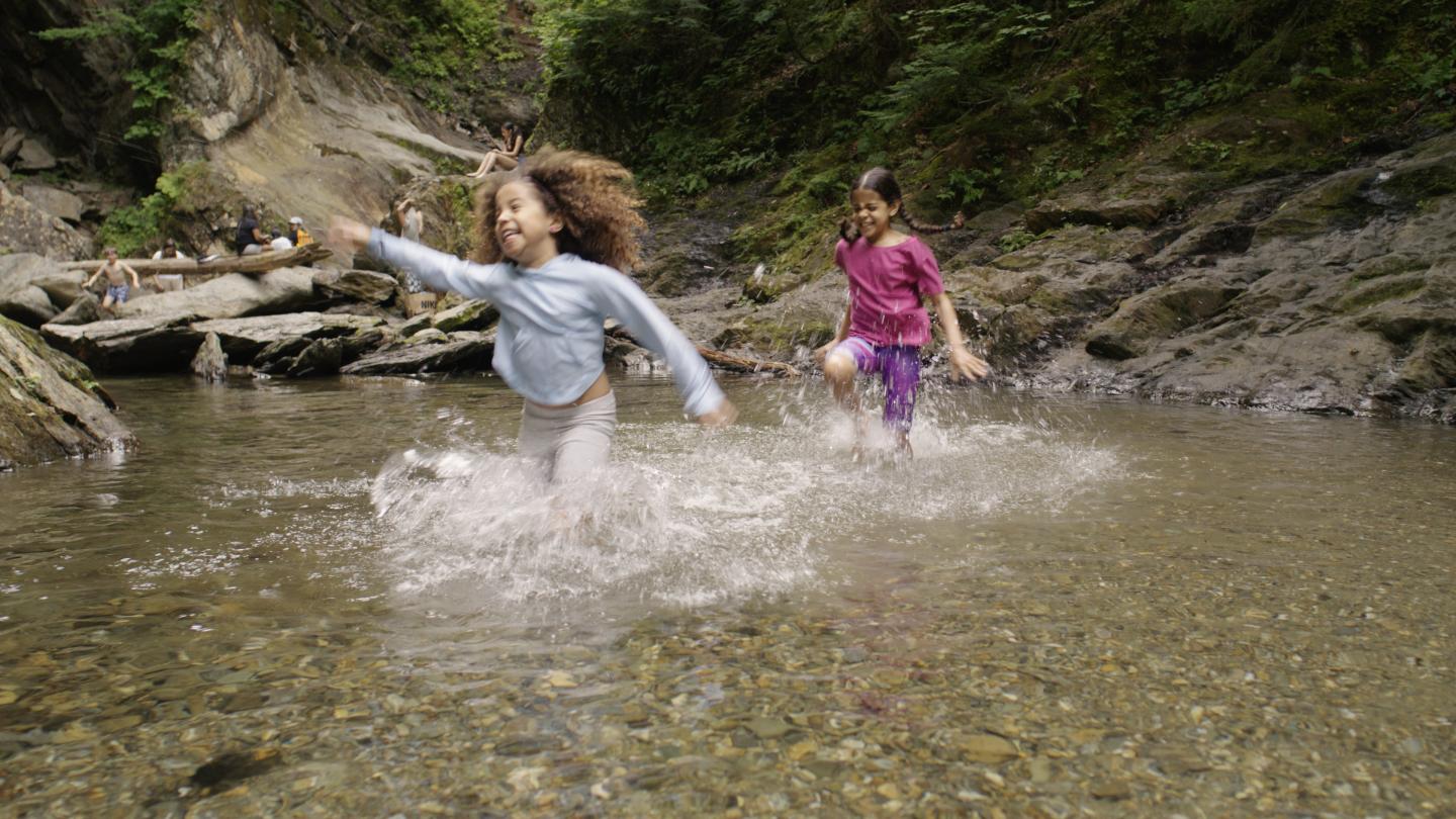 Children joyfully splashing through a shallow, clear stream in a forested area.