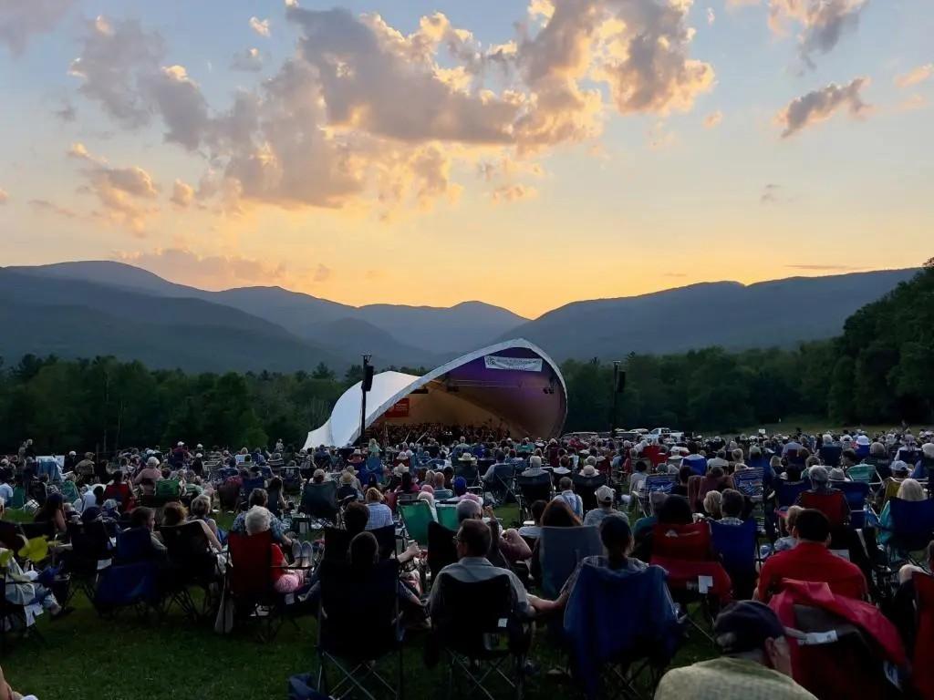Outdoor concert sunset, audience seated on grass, mountains in background.