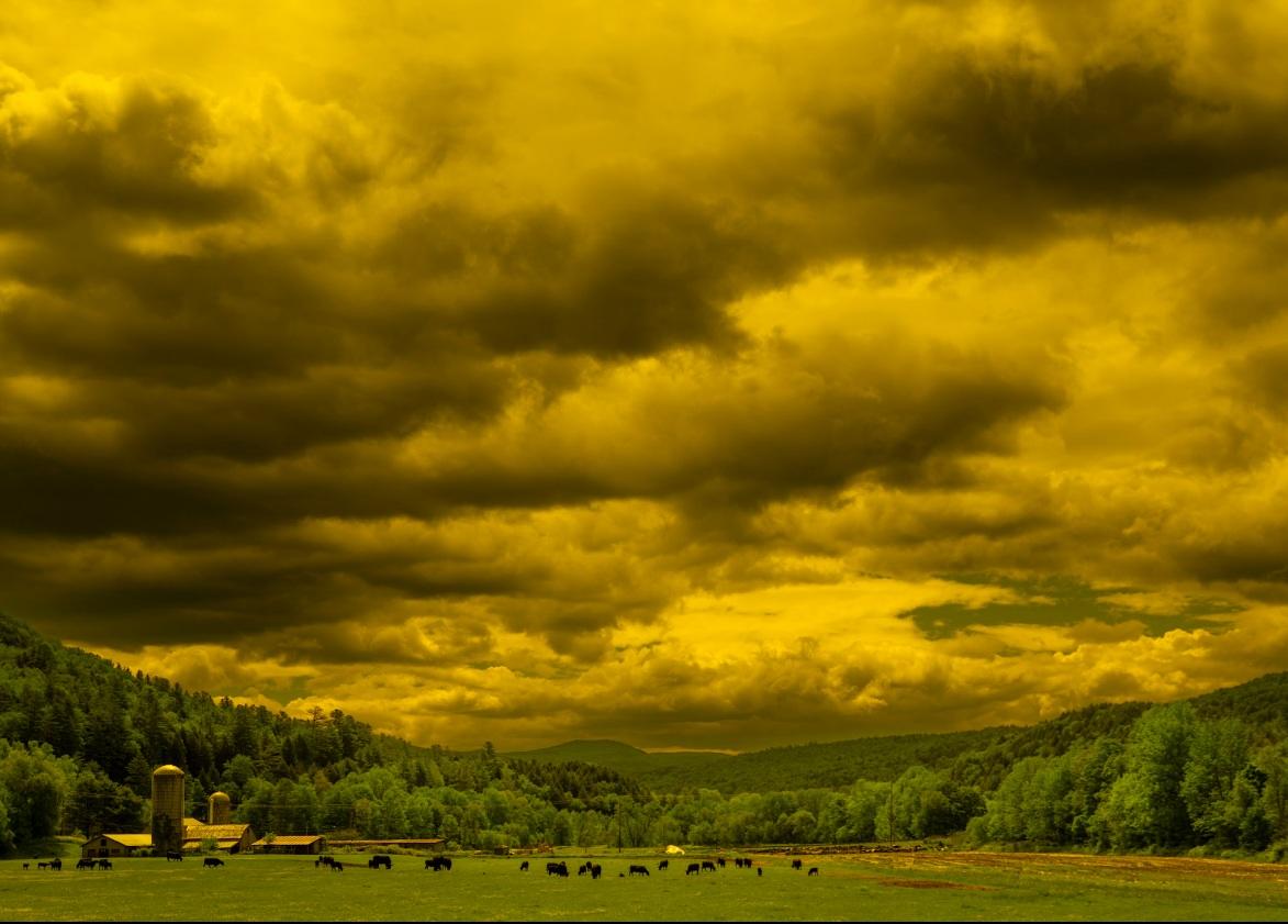 Dramatic cloudy sky over a green valley with trees and distant hills.