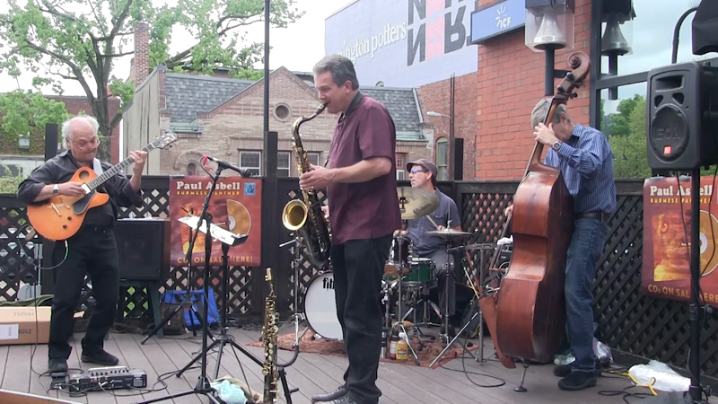 Jazz band performing outdoors with guitar, saxophone, drums, and double bass.