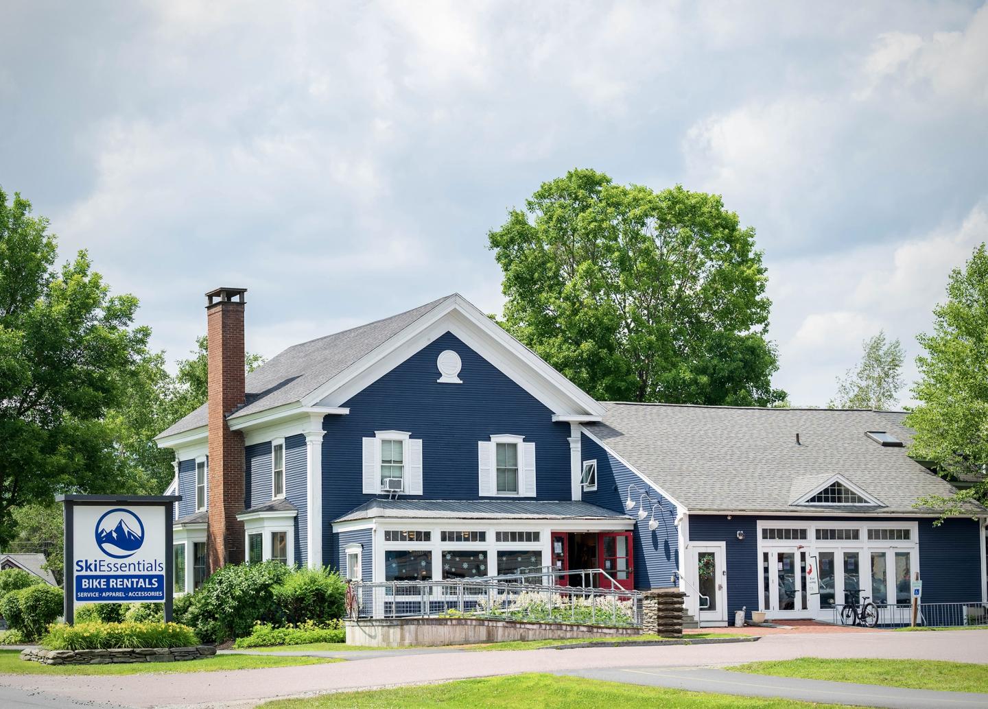 Large blue house with white trim, surrounded by trees and a cloudy sky.