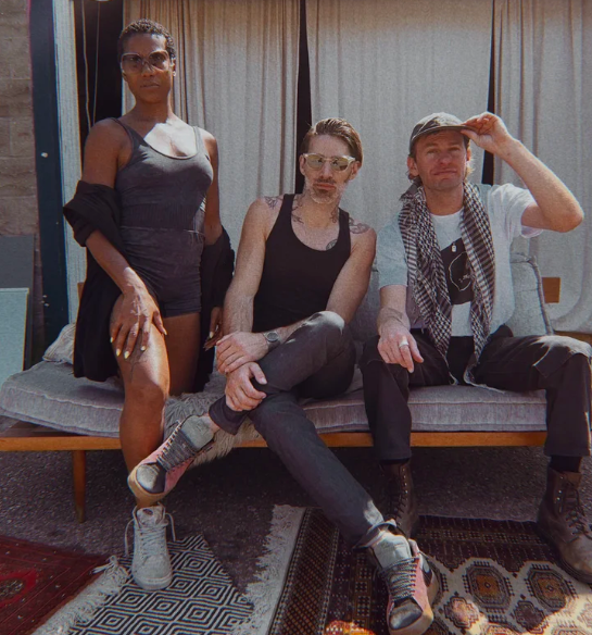 Three people posing outdoors on a couch, wearing casual attire and sunglasses.