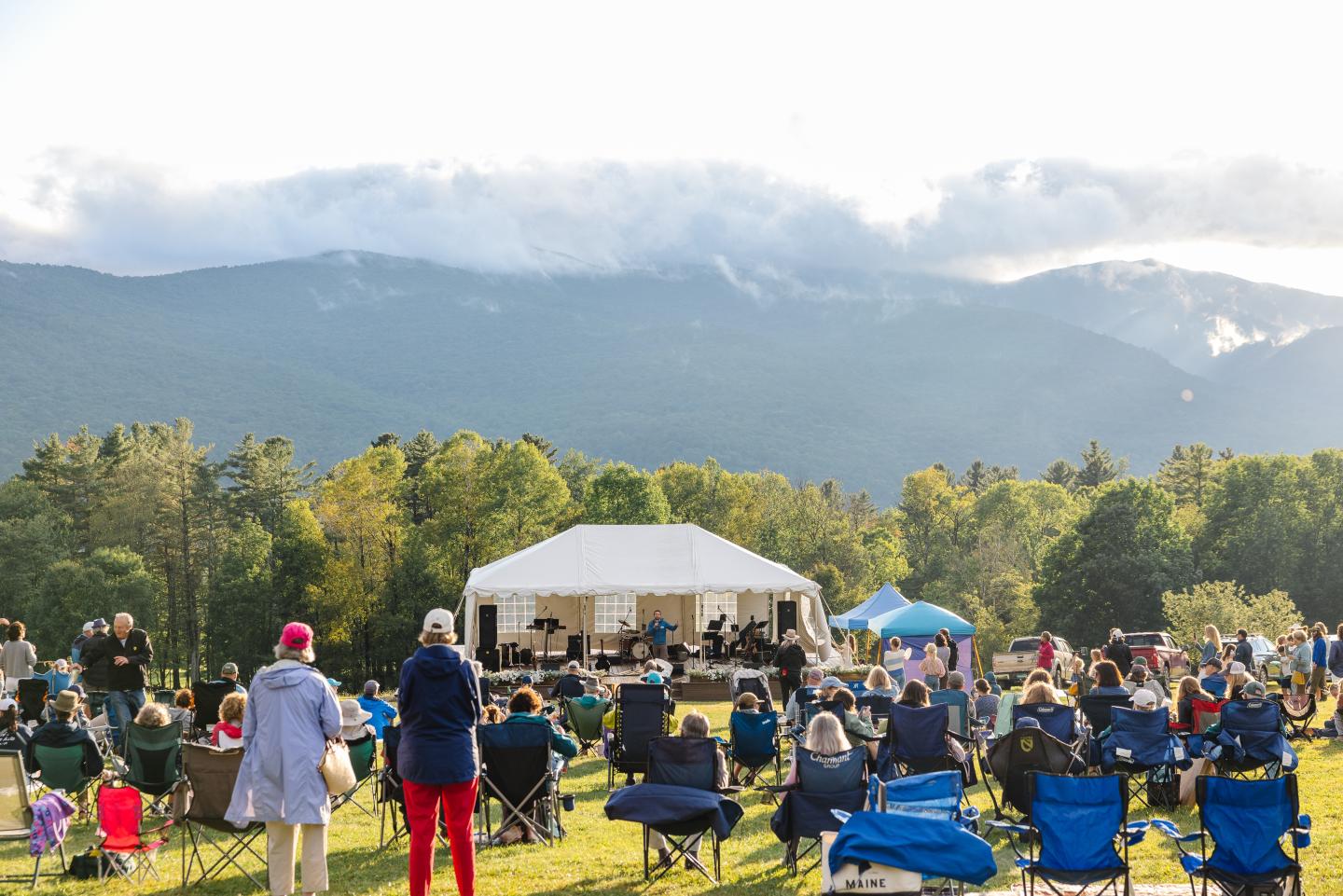 Outdoor concert with people seated on grass, mountains in the background.