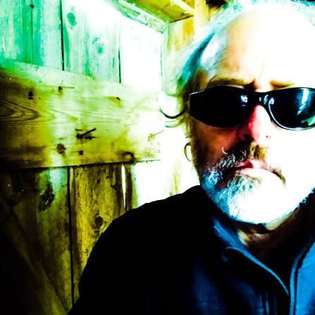 Man with sunglasses and a beard, standing in front of a wooden wall.