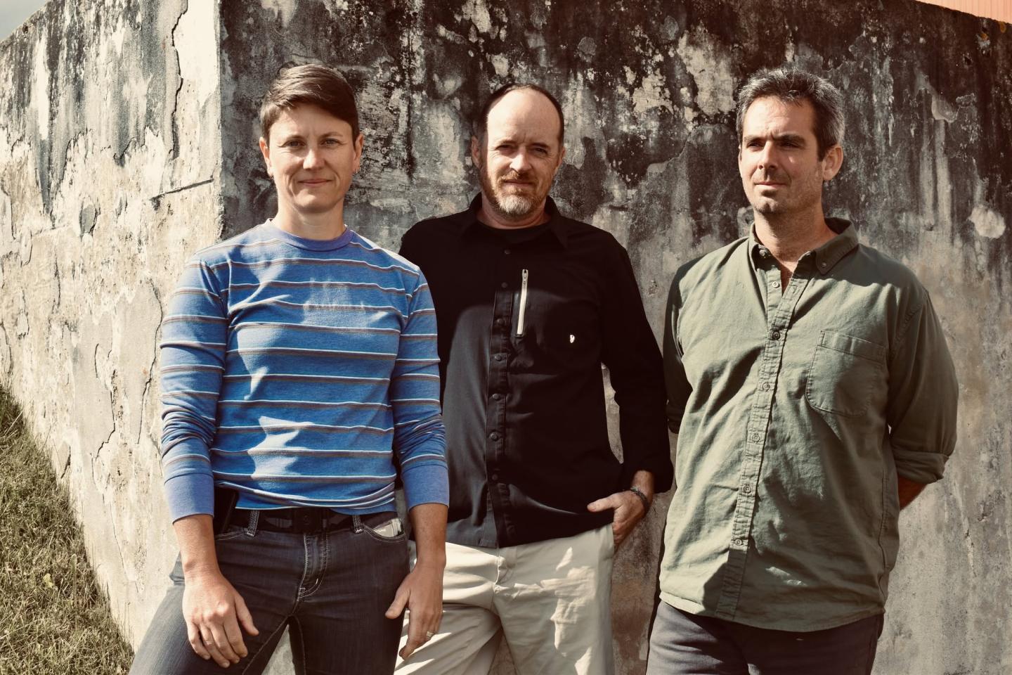 Three people standing outdoors against a textured wall.
