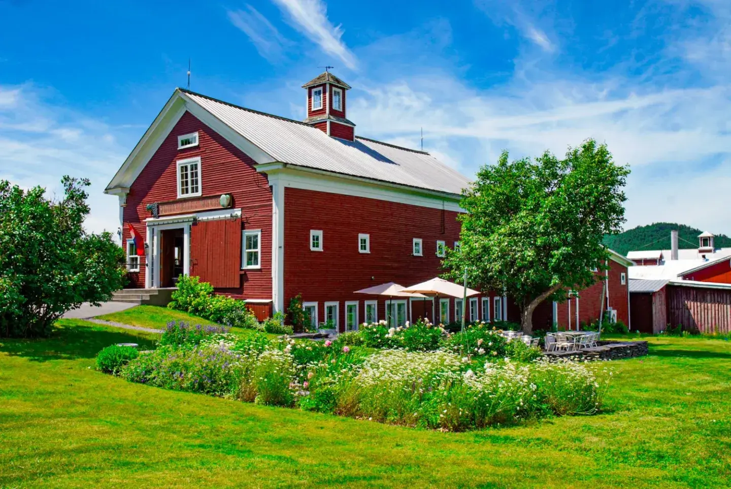 Red barn with white trim, surrounded by green lawn and colorful gardens under a blue sky.