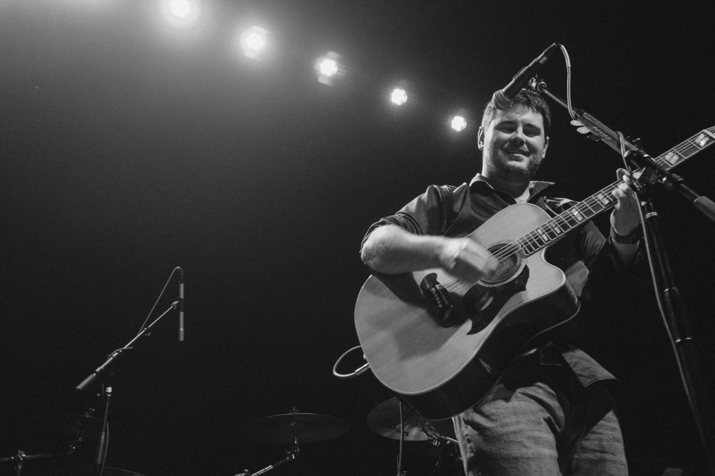 Guitarist performing on stage under bright lights, smiling. Black and white photo.