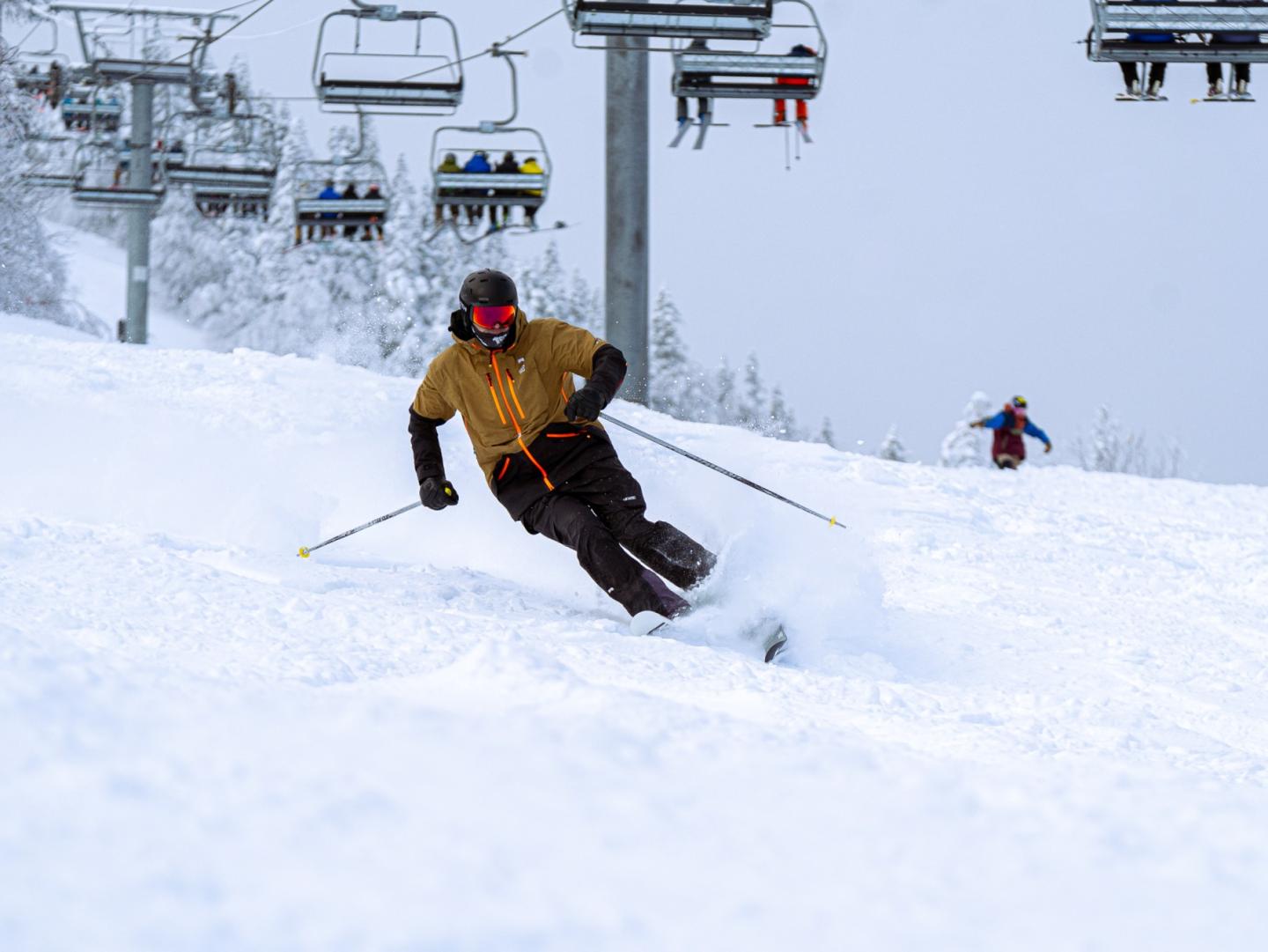 Skier in brown jacket navigating snowy slope, with chairlift in background.