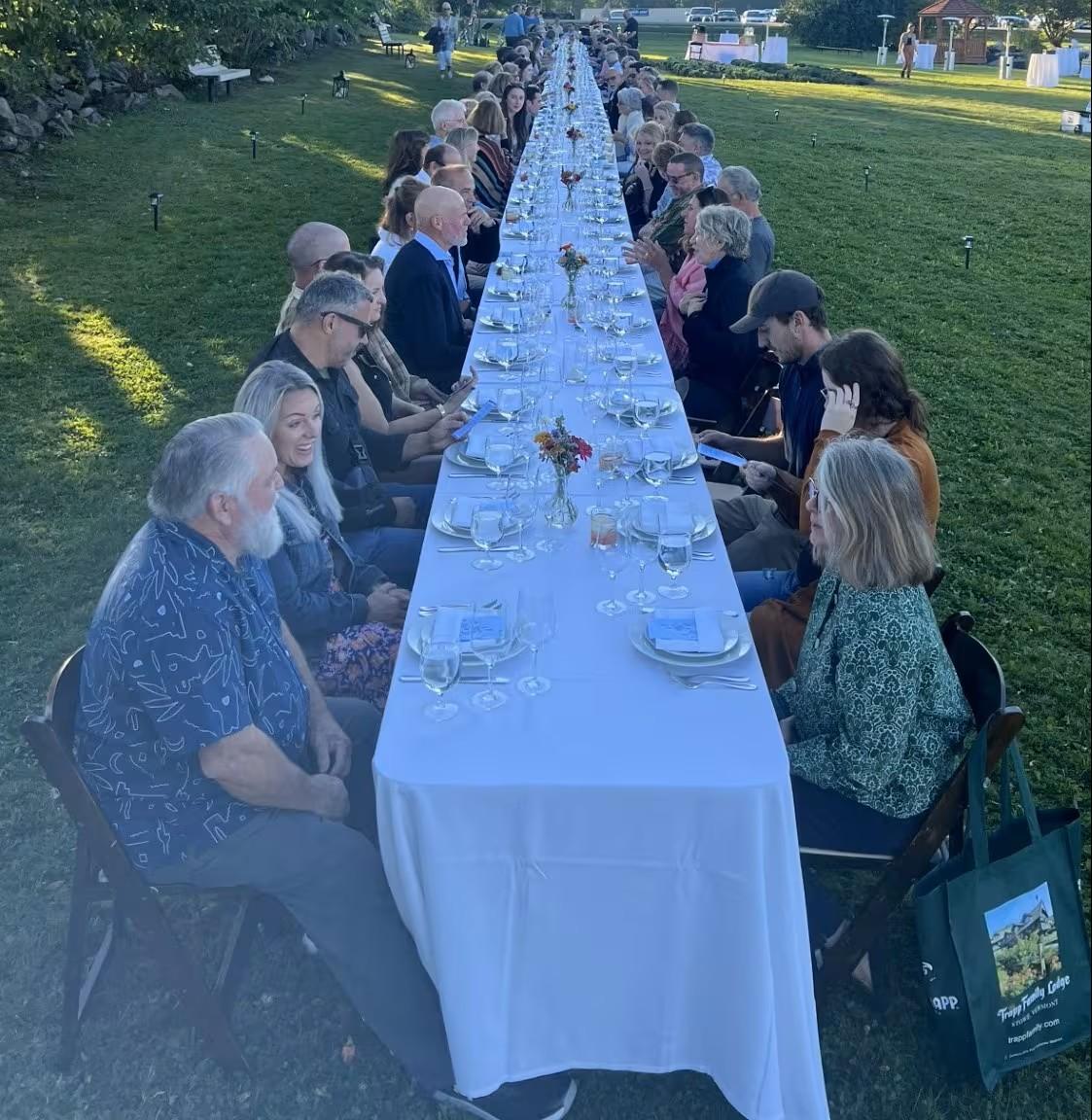 A long outdoor banquet table set with plates and glasses, guests seated along both sides.