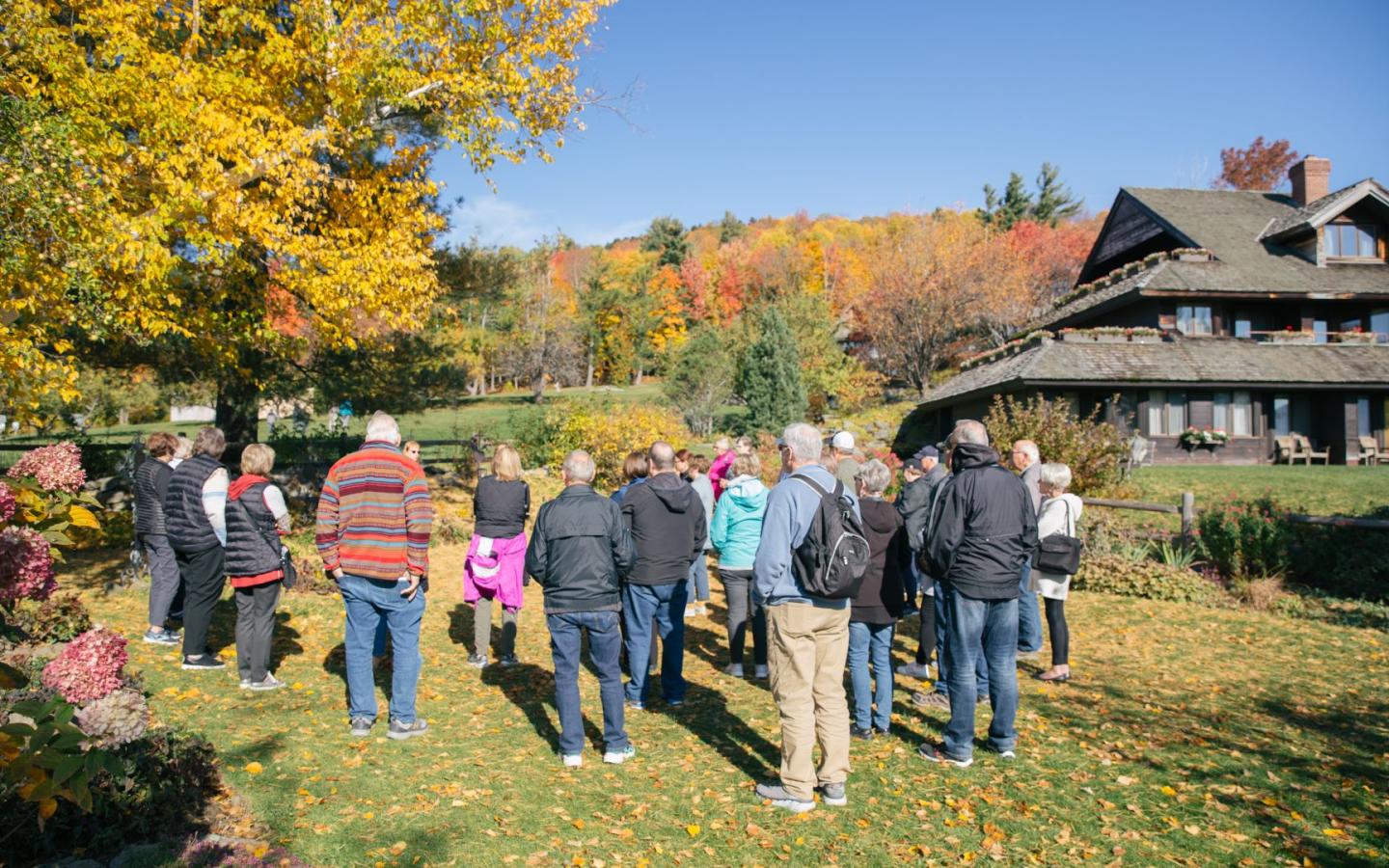 Visitors gather on a lawn near a rustic lodge amid autumn foliage.