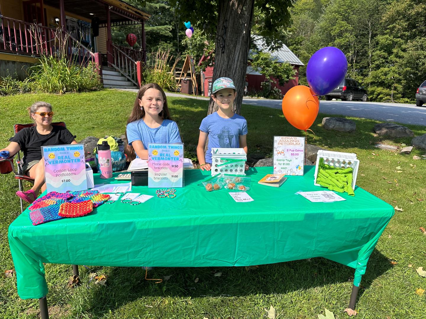 Young people at a green table outdoors, displaying crafts and items with colorful balloons.