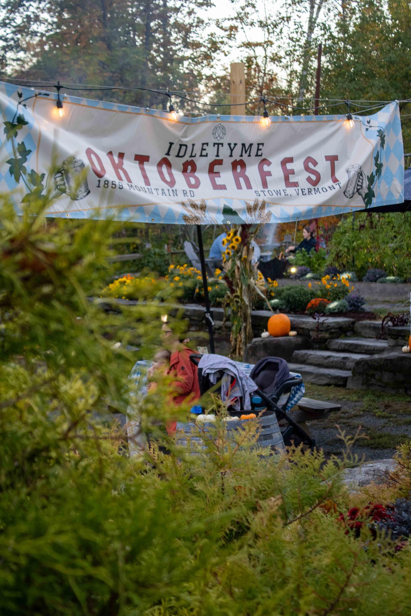 Outdoor festival banner reading “Welcome Oktoberfest” above people by a pond.