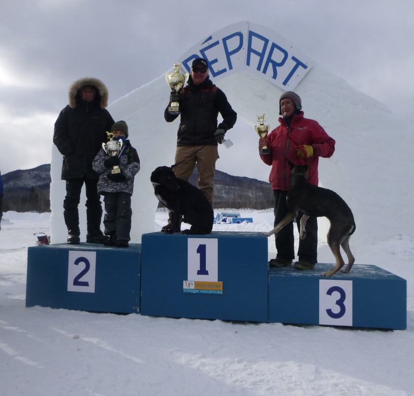 Winners Jim on podium holding trophies, accompanied by two dogs, in snowy setting.