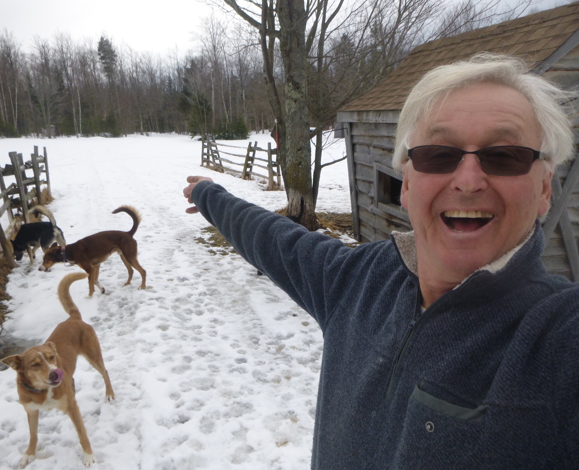 Jim smiling, pointing in snowy yard with sled dogs, near rustic cabin.
